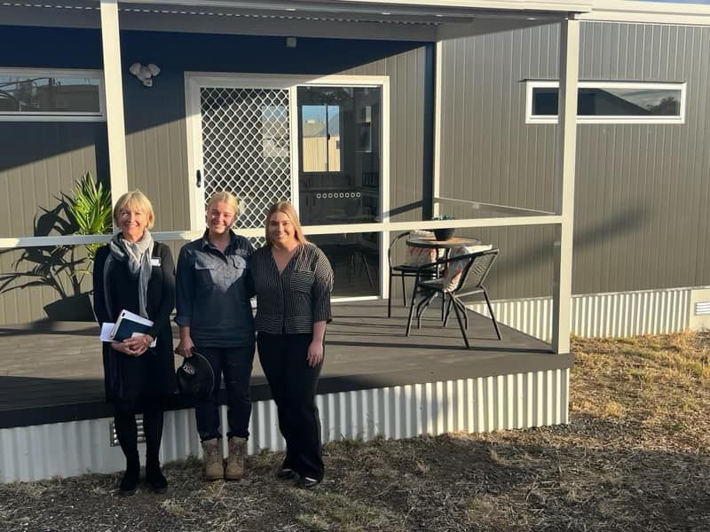 Three people standing on a wooden porch in front of a modern, dark grey modular building.