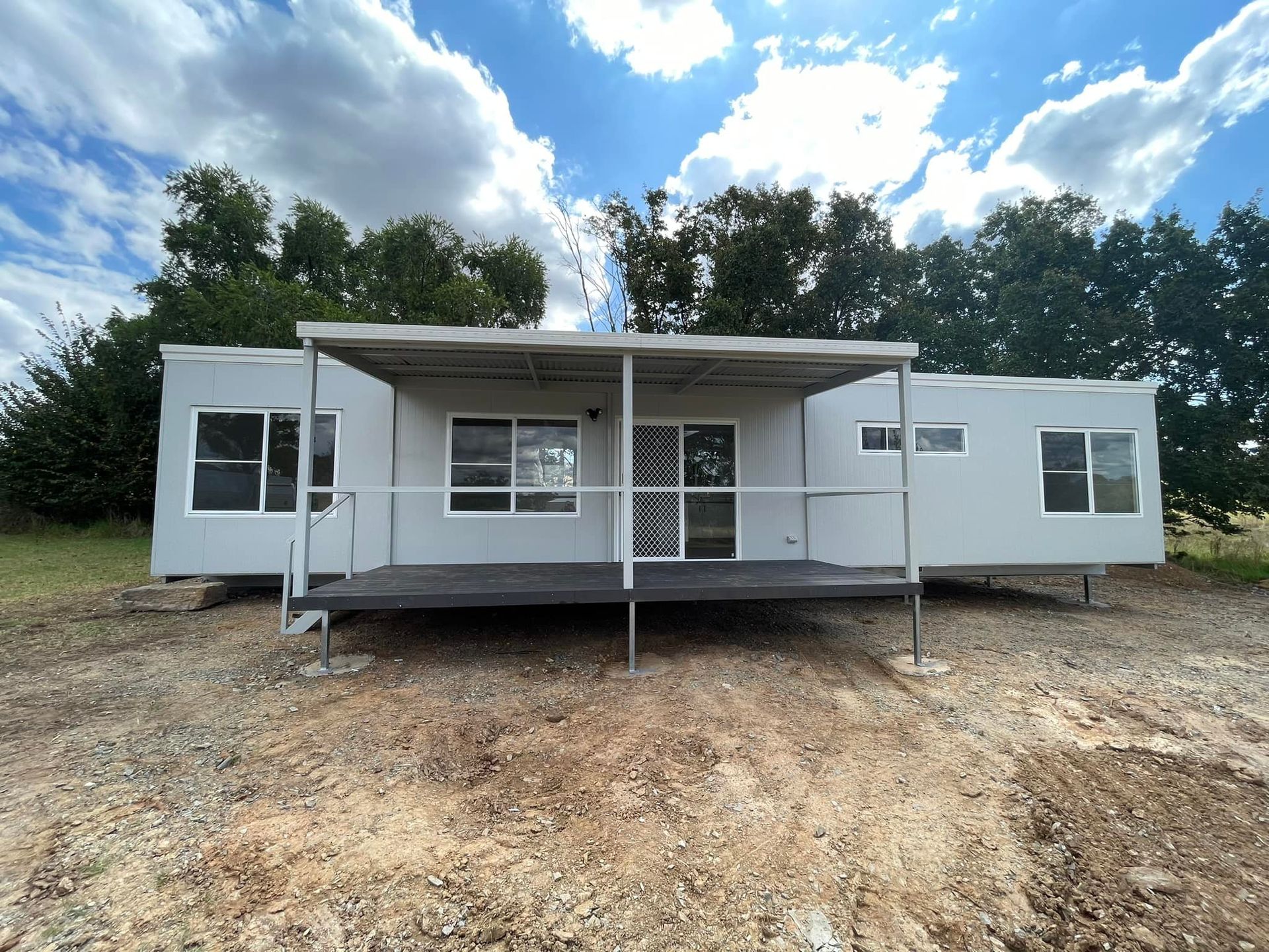 A Light Grey Modular Home With a Dark Deck and Covered Porch Sits on an Undeveloped Dirt — OSM Transportables In Dubbo, NSW