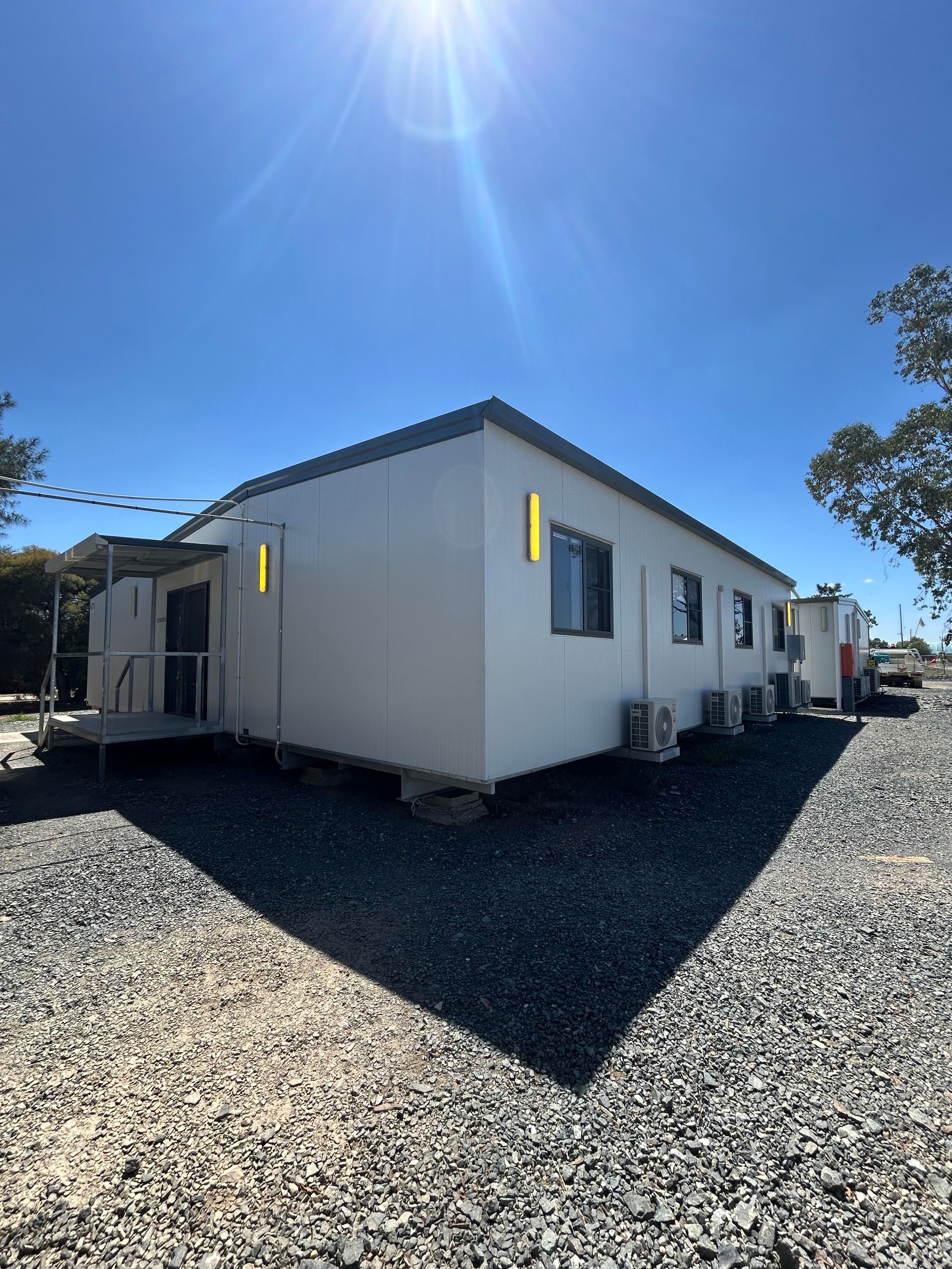 A White, Single-story Modular Building Sits on a Gravel Lot Under a Bright — OSM Transportables In Dubbo, NSW