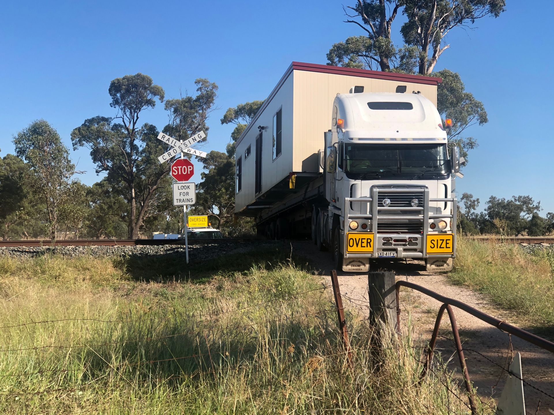 A White Semi-truck Carrying a Large Modular Building Crosses a Railroad Track — OSM Transportables In Dubbo, NSW