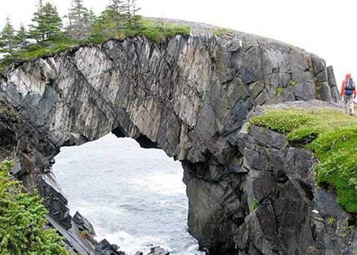 Natural rock archway over ocean, with person hiking on the right side.