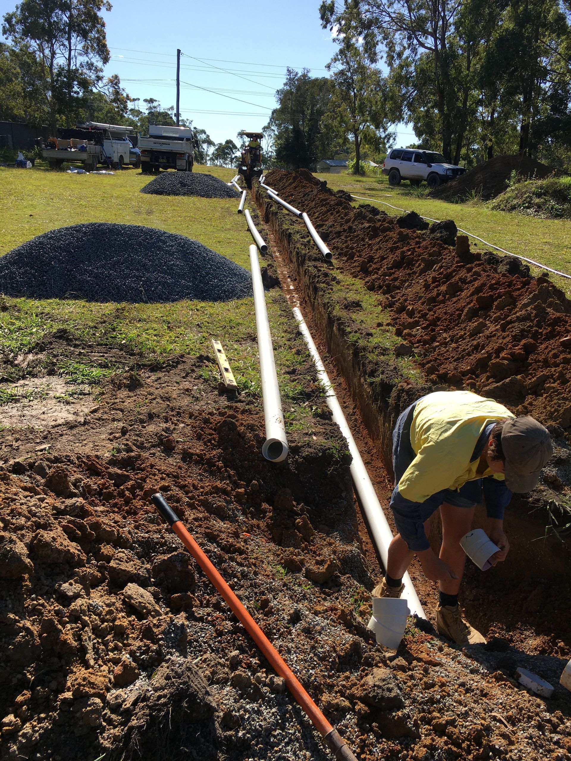 Man installing white pipes in a trench. Construction equipment and gravel pile on a grassy field— VR Marsden Plumbing in Red Rock, NSW