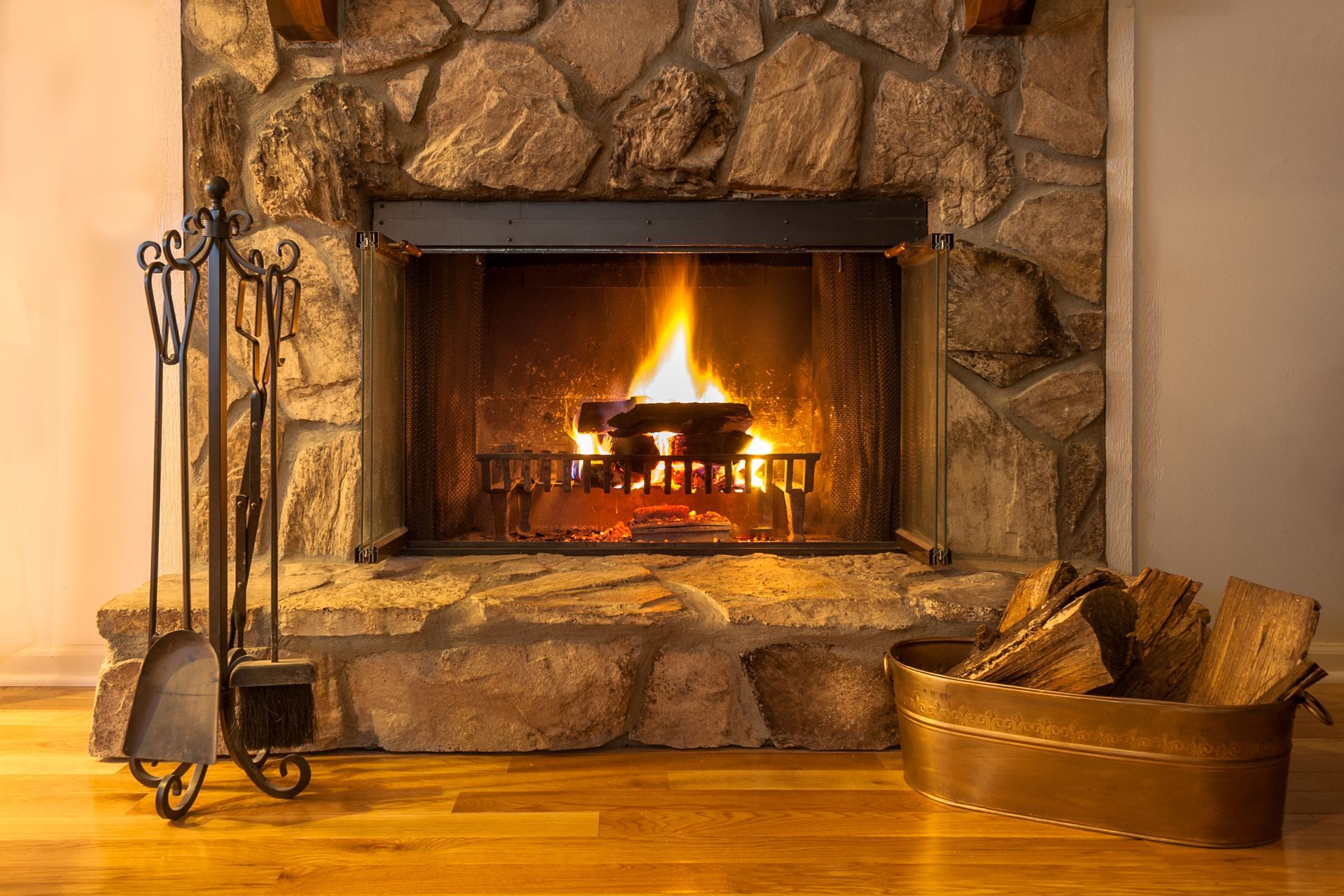 Lit fireplace in a stone hearth with firewood basket and fireplace tools on a wooden floor