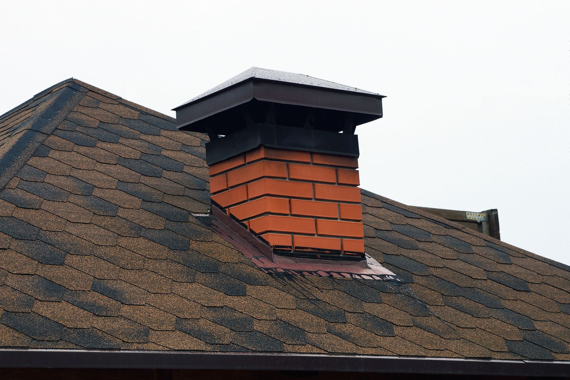Close-up of a rooftop chimney with red brick and a dark metal cap on asphalt shingles