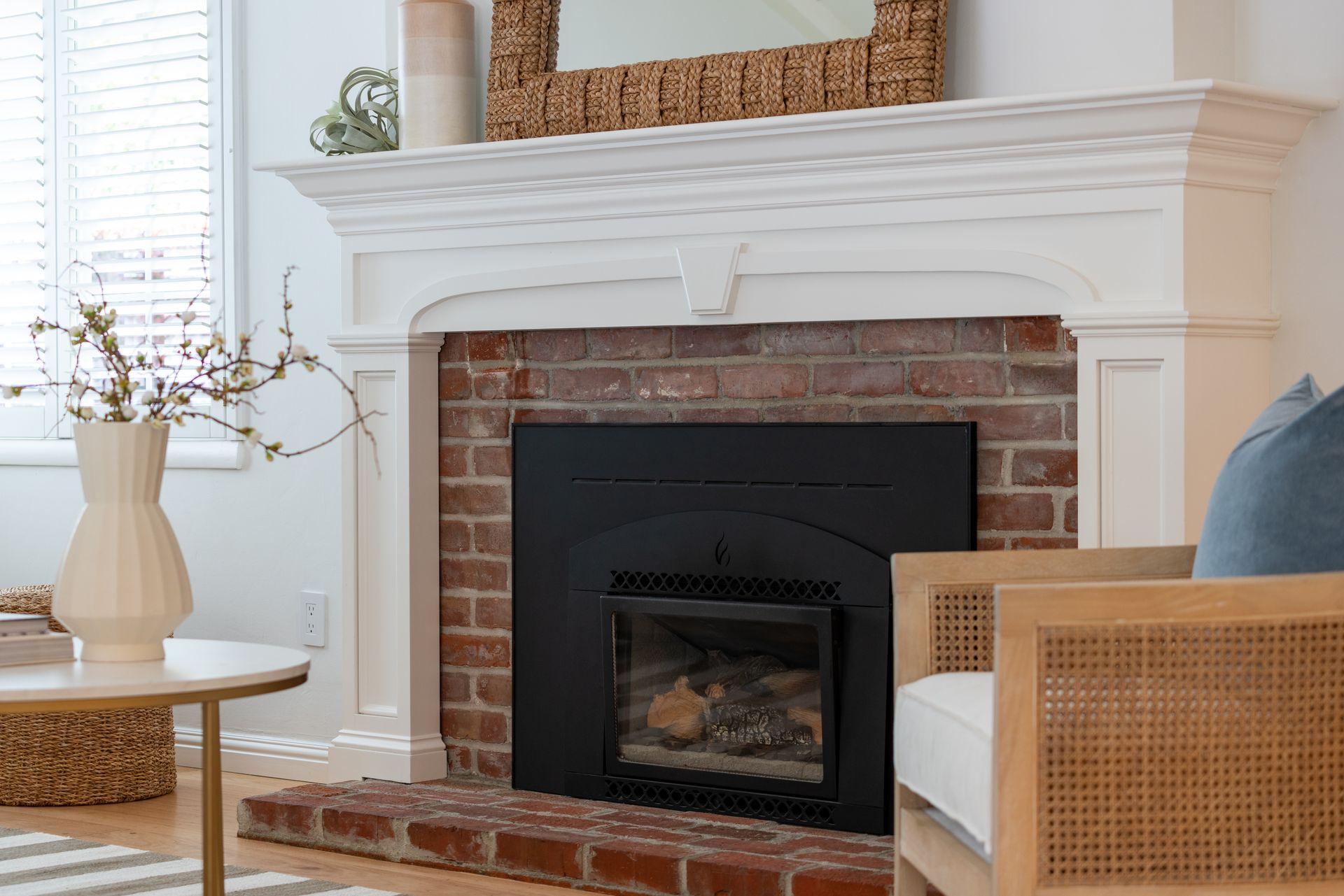 Bright living room fireplace with white mantel, brick surround, and wicker chair beside a round table