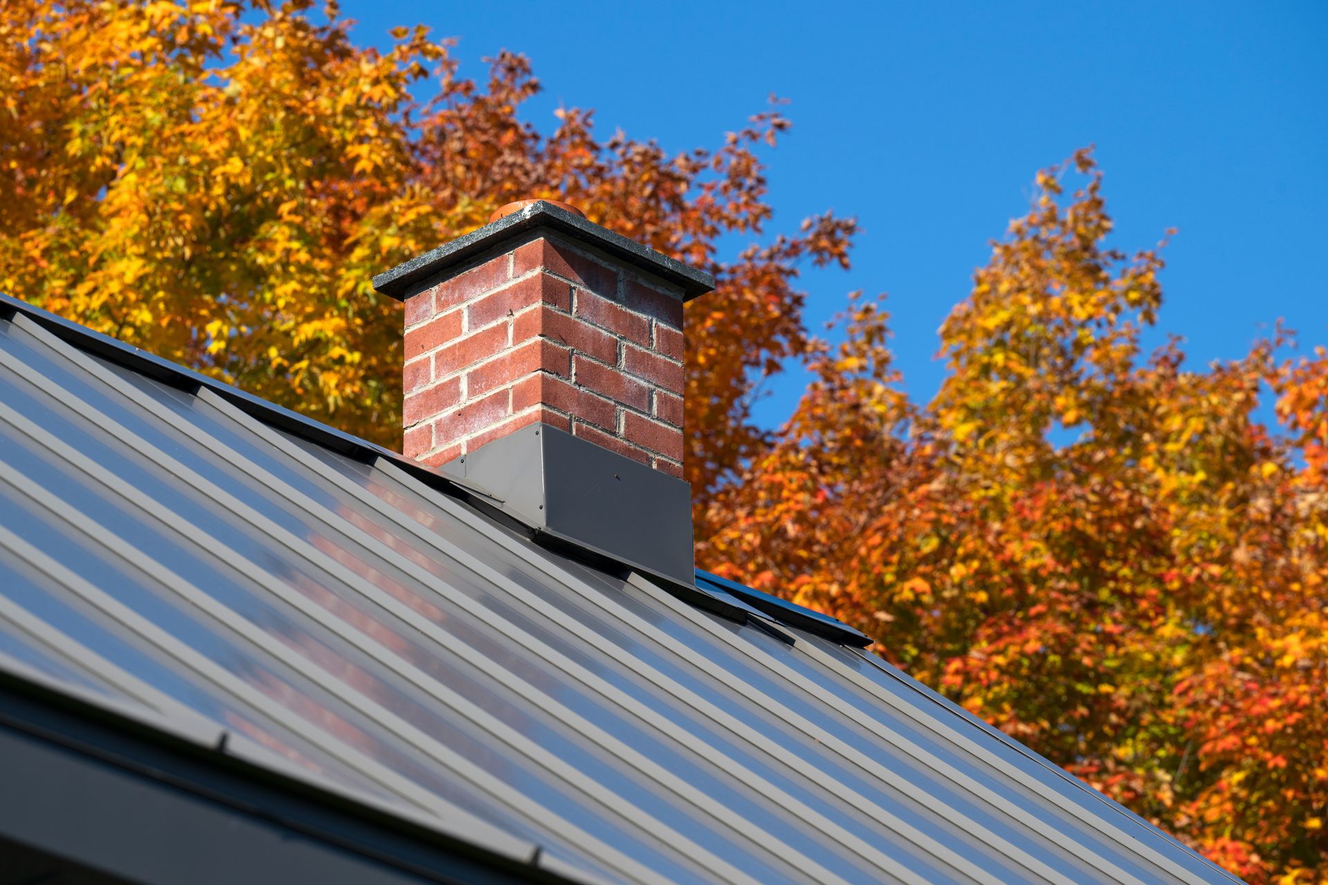 Brick chimney on a metal roof with bright autumn trees and blue sky in the background