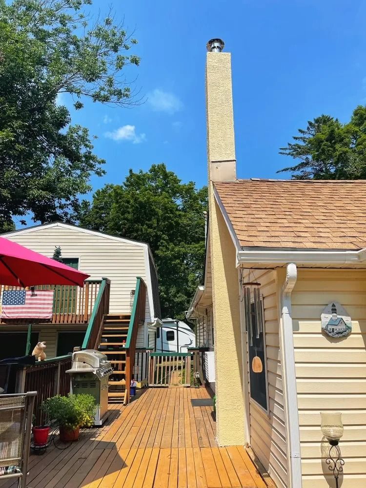 Narrow wooden deck between beige houses with stairs, potted plants, and a tall chimney under a clear blue sky