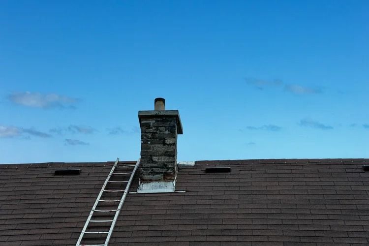 Roof with a ladder leaning against a brick chimney under a blue sky.