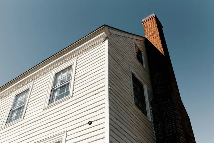 Corner of a white house with windows and a brick chimney against a blue sky