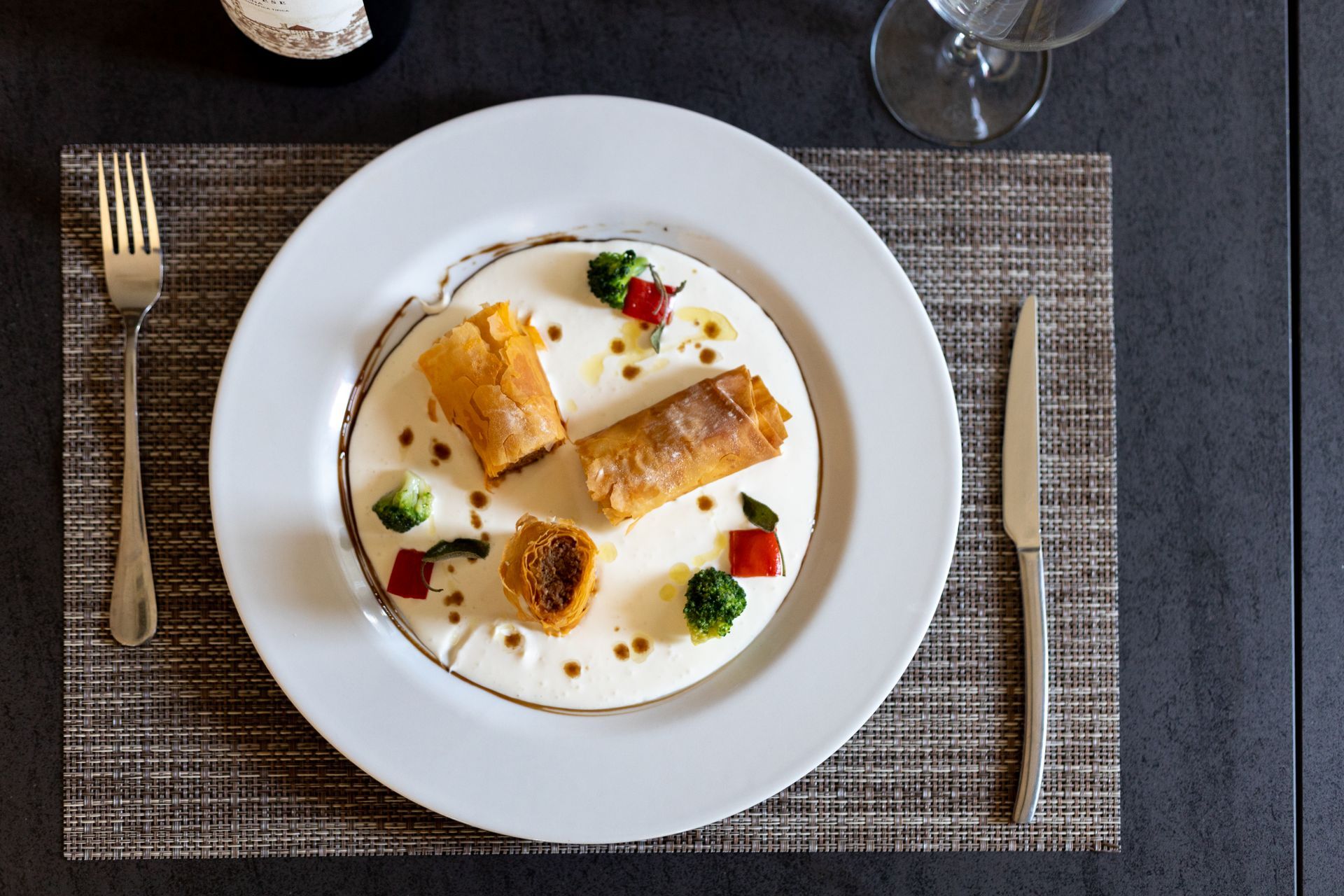Dessert gourmet servito su un piatto bianco con frutti di bosco e panna, adagiato su una tovaglietta intrecciata.