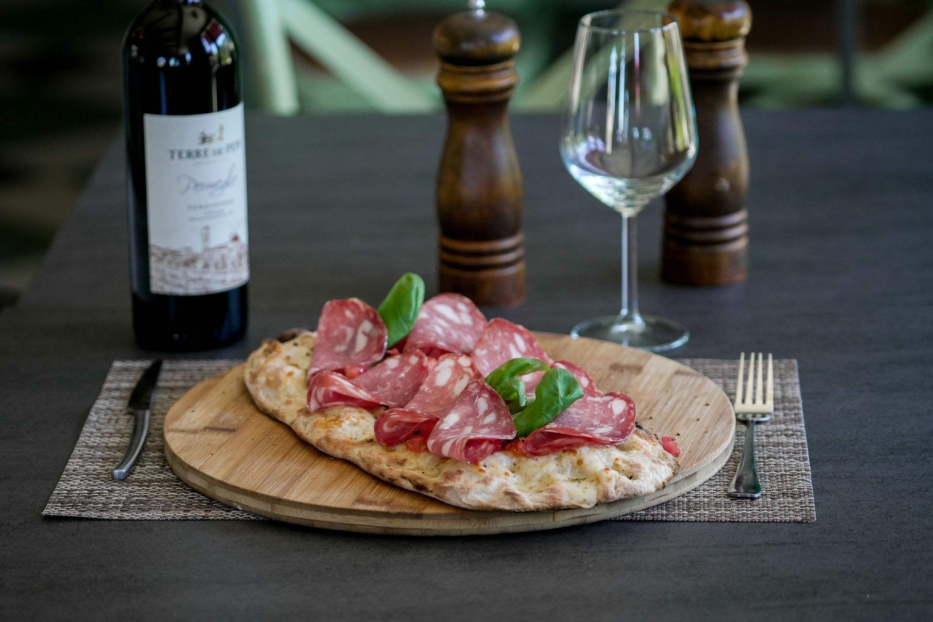 Una focaccia con salumi e basilico su un piatto, accanto a un bicchiere da vino, un bicchiere da vino e un macinapepe su un tavolo scuro.