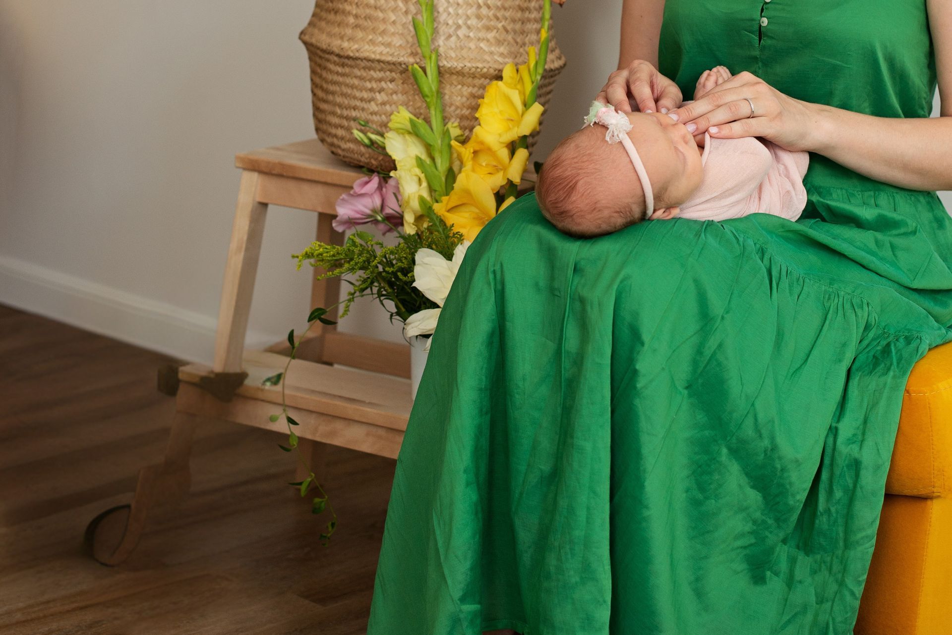 Woman in green dress holds a newborn wearing a pink onesie and headband. Flowers and a basket sit nearby.