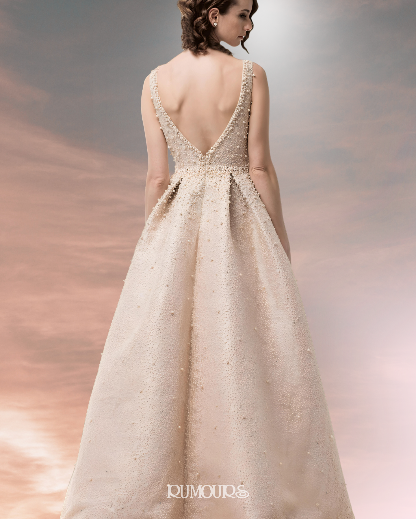 Woman in a beaded, open-back gown, standing with back to the viewer against a soft, colorful background.
