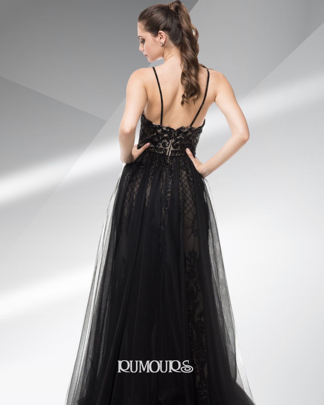 Woman in a black evening gown, hands on hips, back to the camera, in a studio setting.