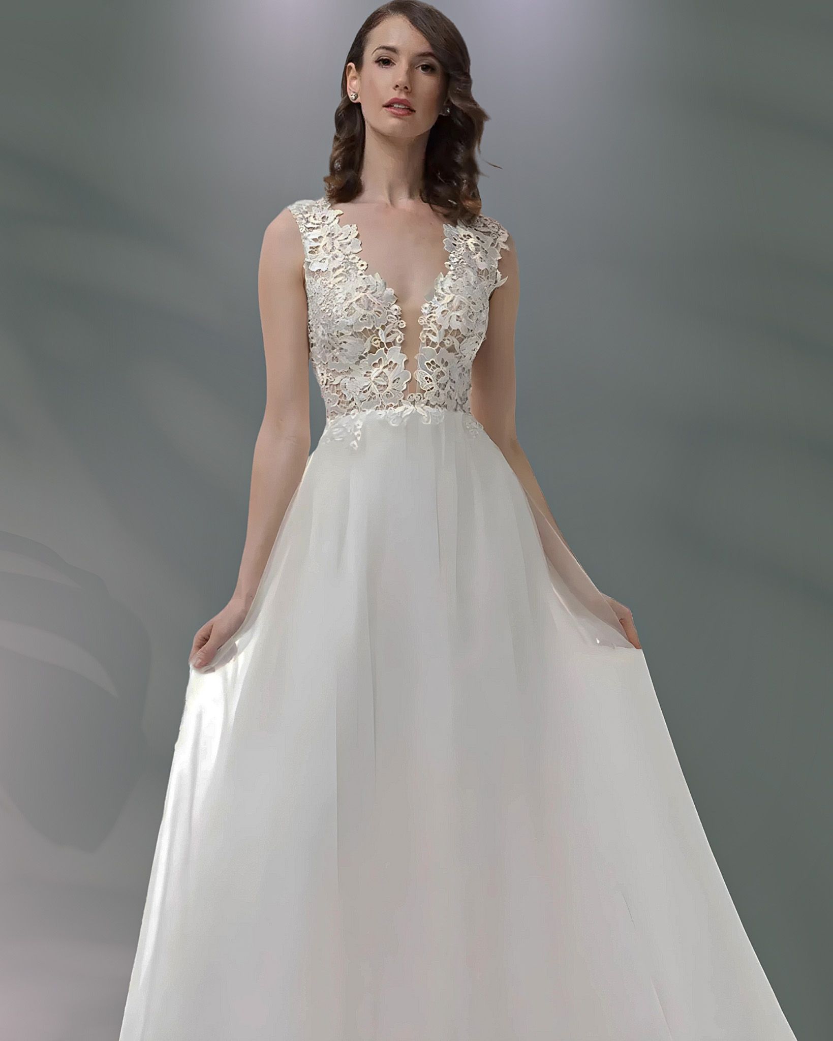 Woman wearing a white, sleeveless wedding gown with a deep V-neck and a flowing skirt, standing in a studio.