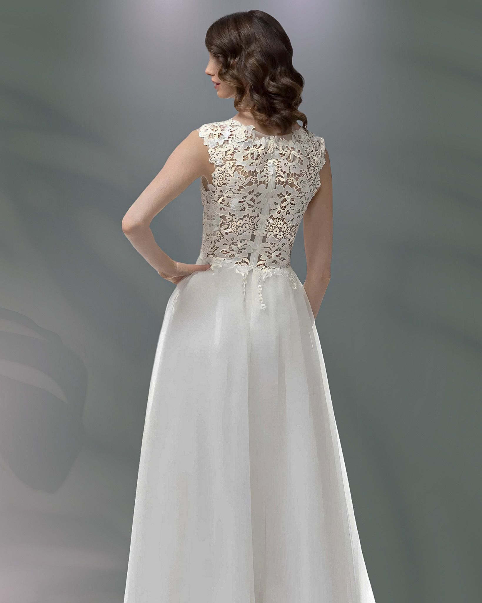 Woman in a white wedding dress, back view, standing indoors, hands on hips.