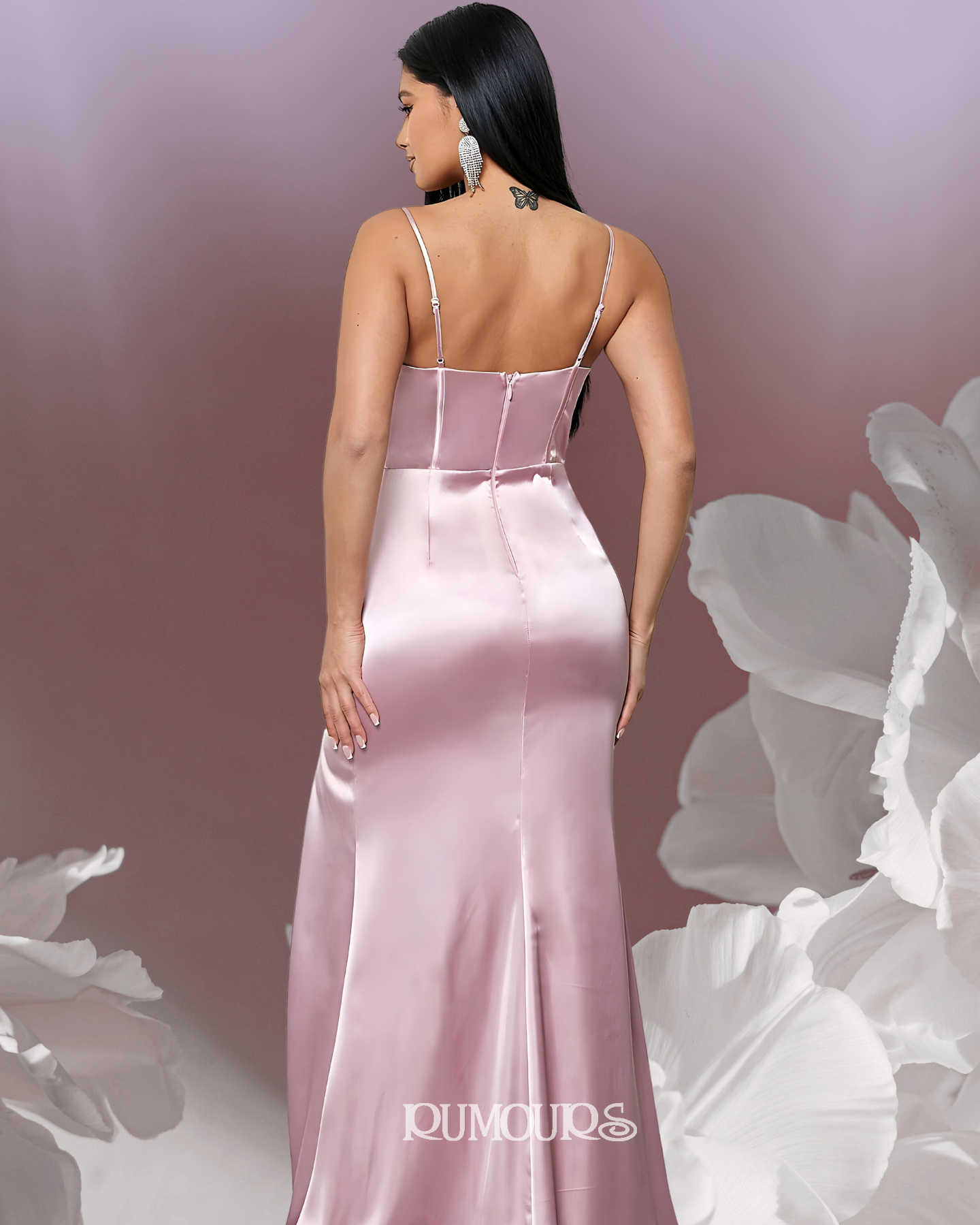 Woman in a flowing pink satin gown, standing with her back to the camera, next to large white floral elements.
