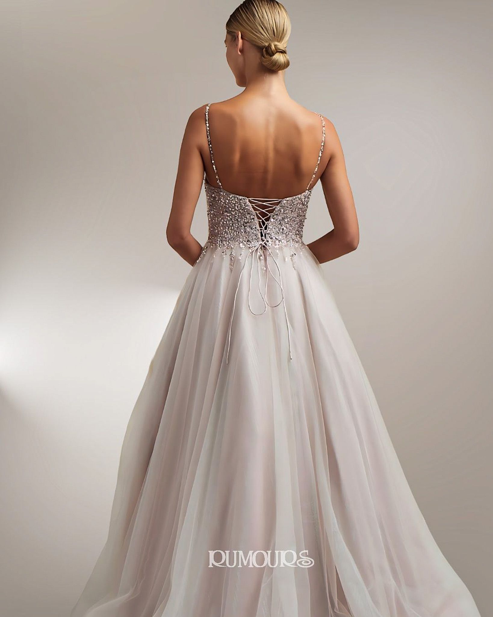Back view of a woman in a light pink embellished gown with spaghetti straps, standing against a neutral background.