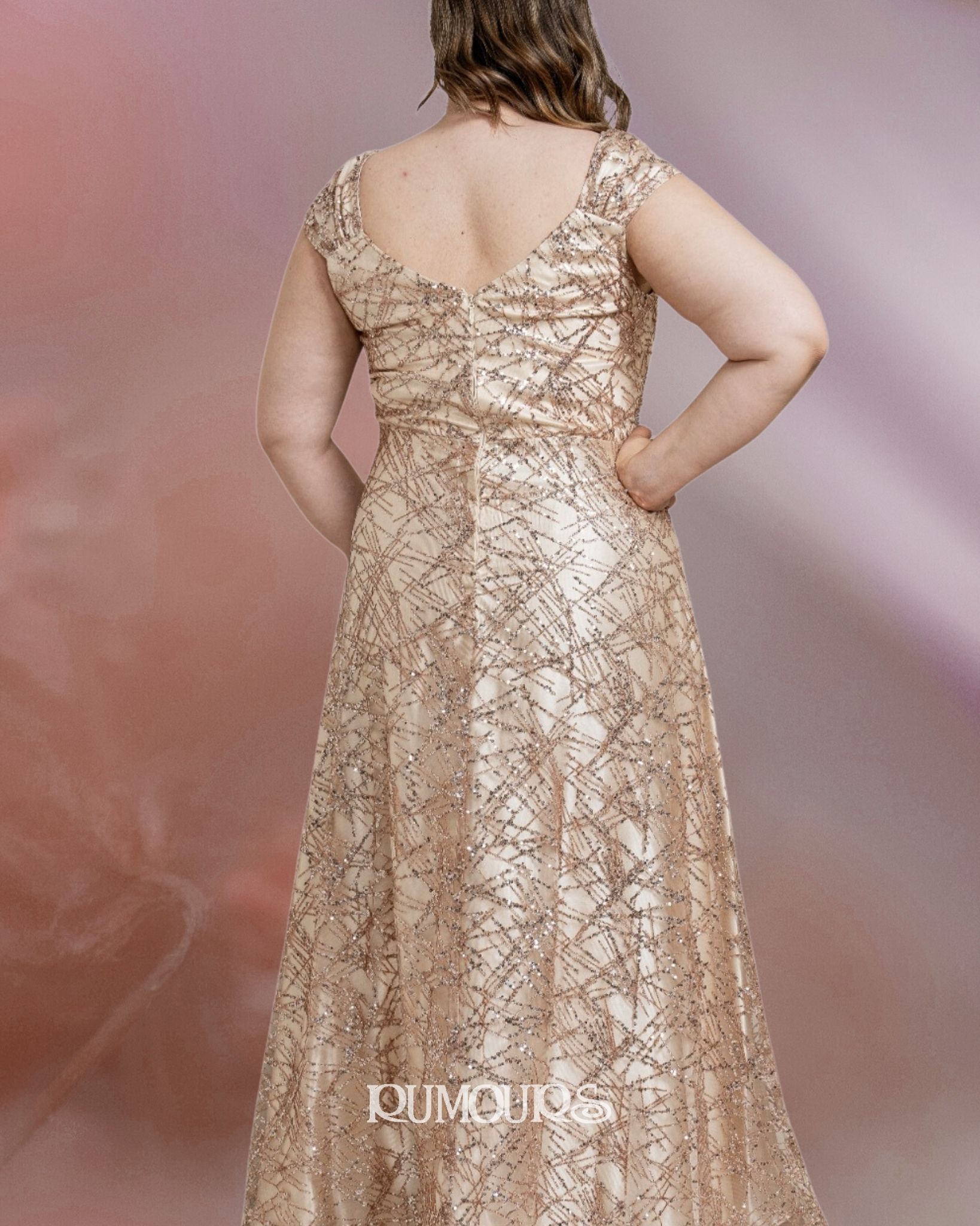 Woman in a gold sequined formal gown, hands on hips, standing in front of a soft pink backdrop.