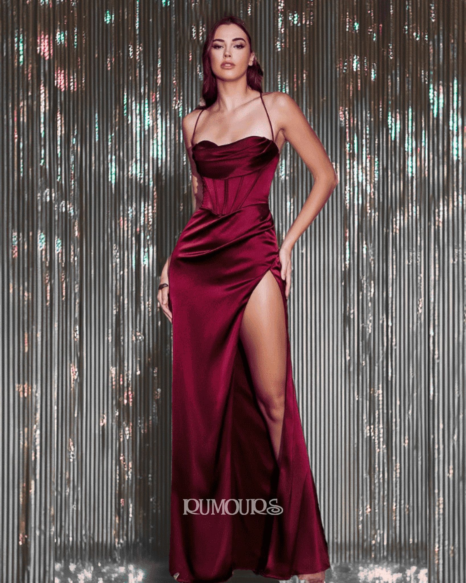 Woman in a burgundy satin gown with a high slit, posing in front of a silver, textured backdrop.