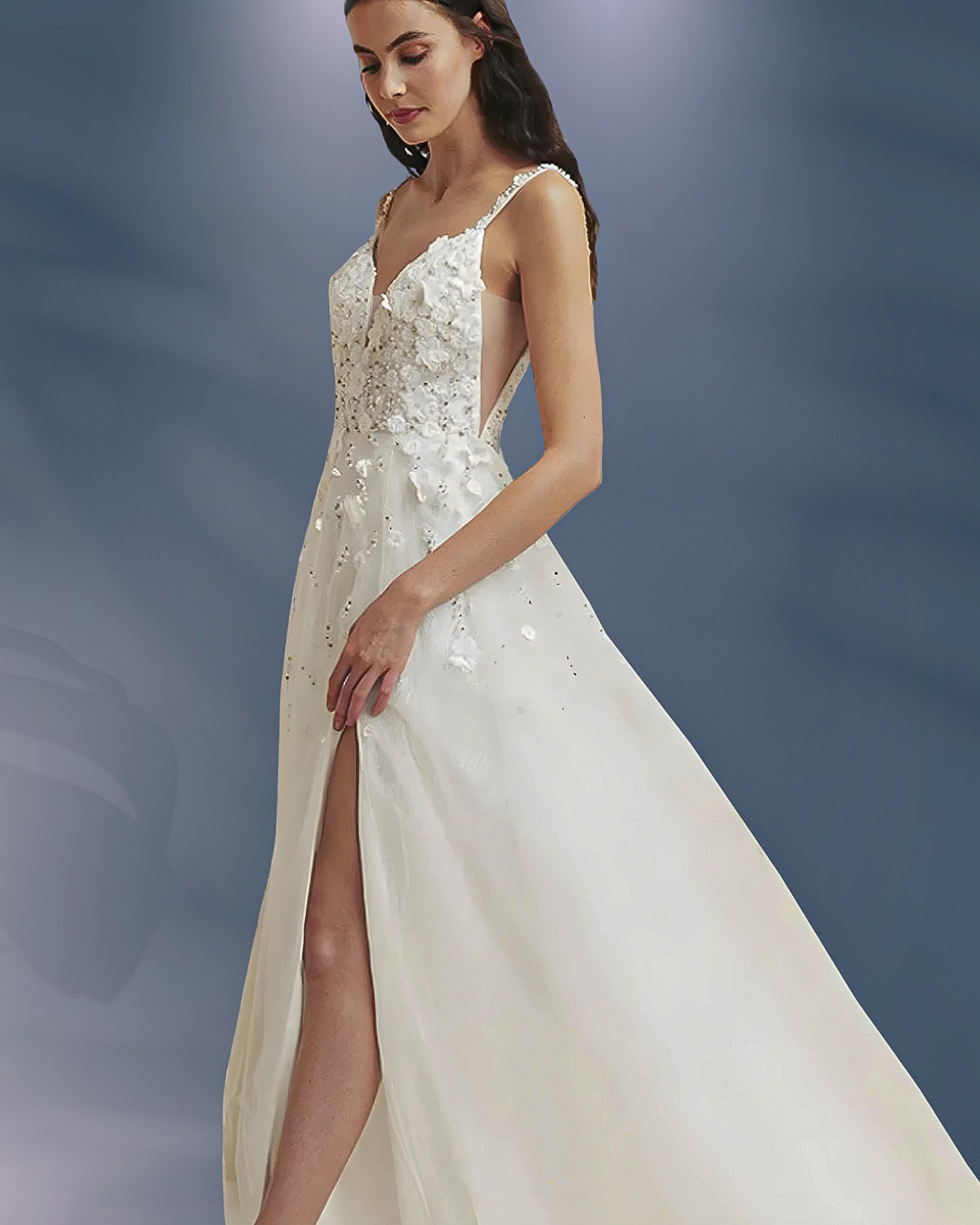 Woman in white wedding dress with a high slit, adorned bodice, and a flowing skirt, set against a blue backdrop.