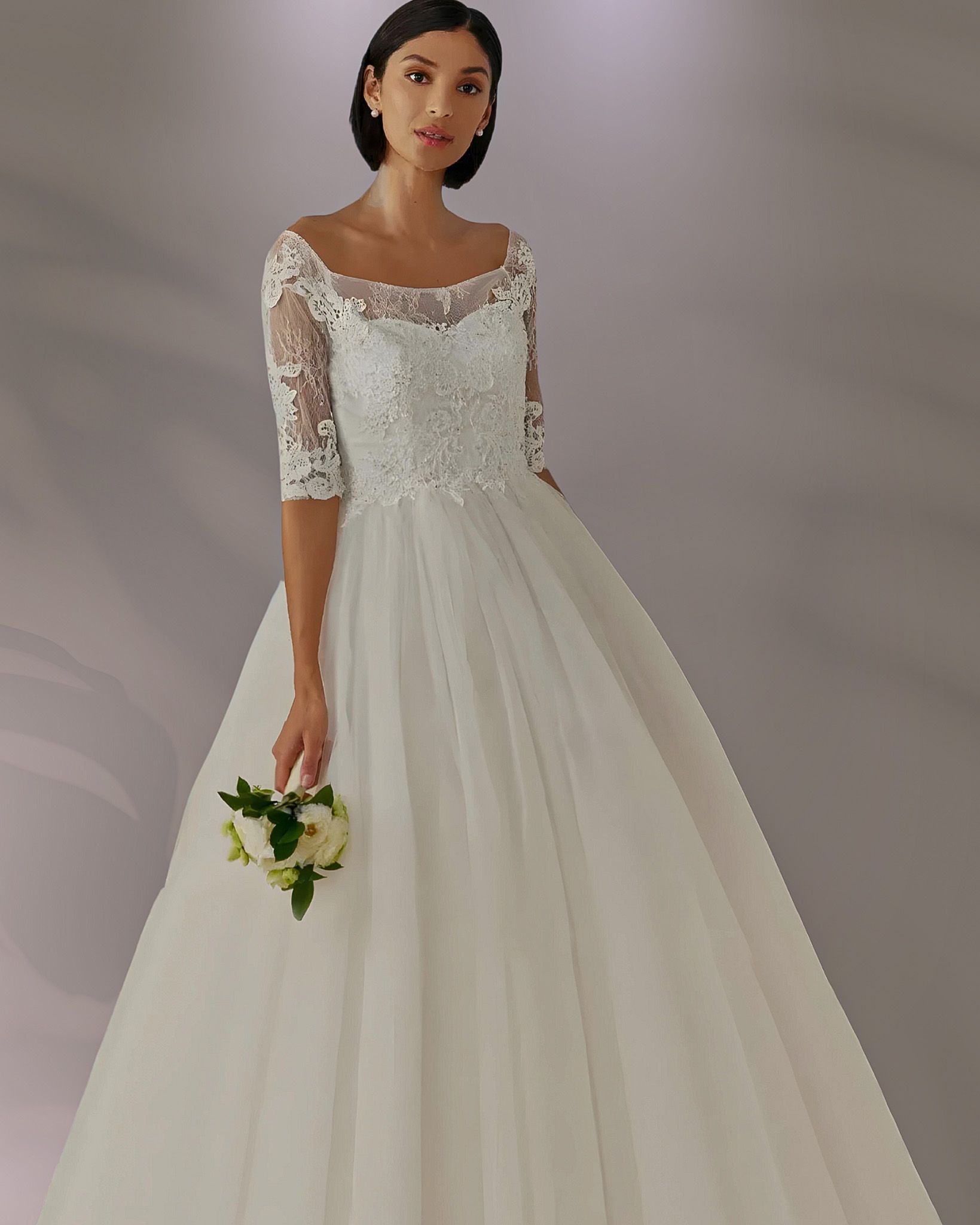 Woman in a white wedding gown, holding a bouquet. She's standing in front of a light-colored background.