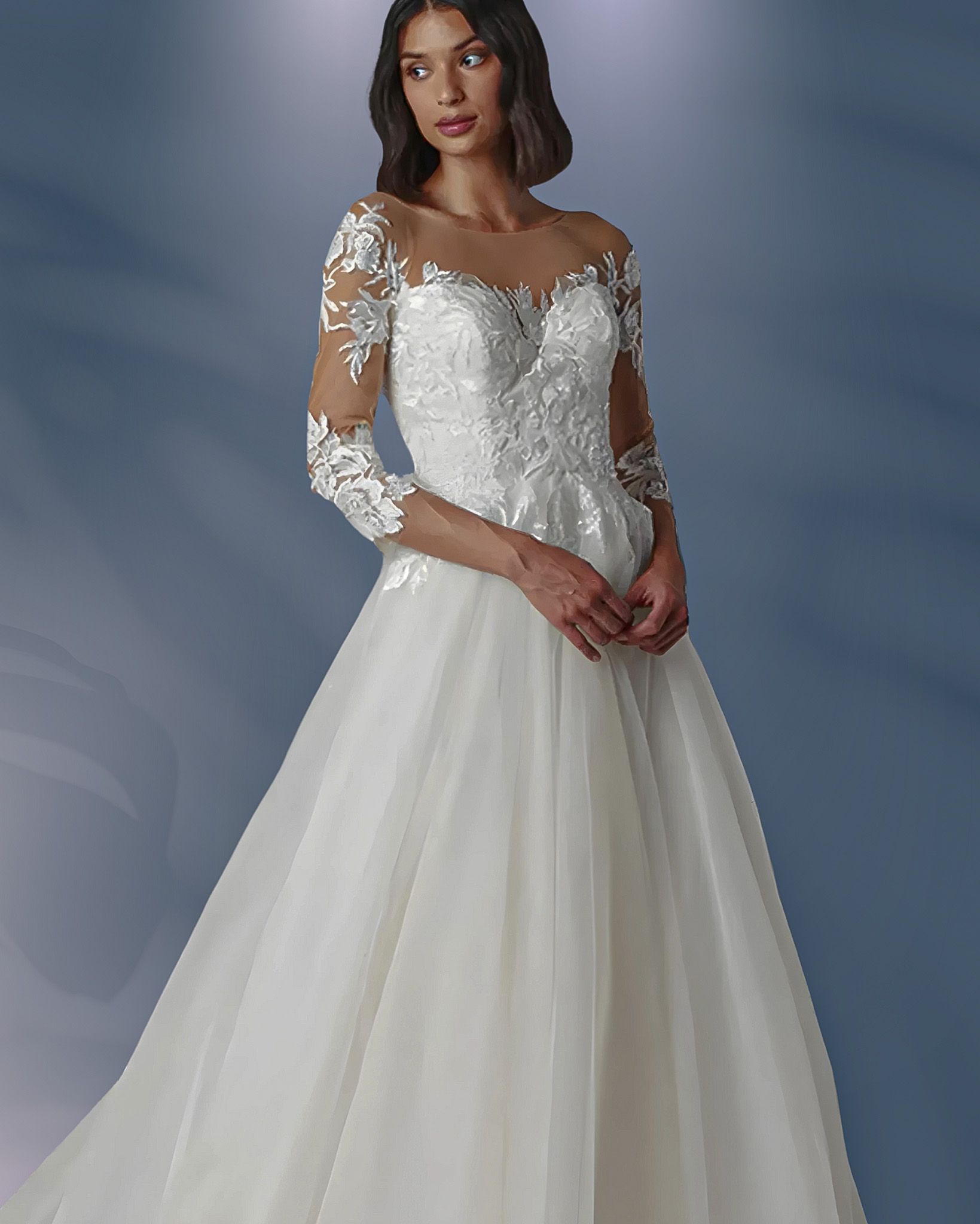 Woman in a white wedding gown with lace sleeves and a tulle skirt, posing in front of a blue background.