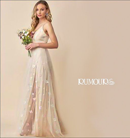 Woman in a cream dress with floral accents, holding flowers, standing near a wall.