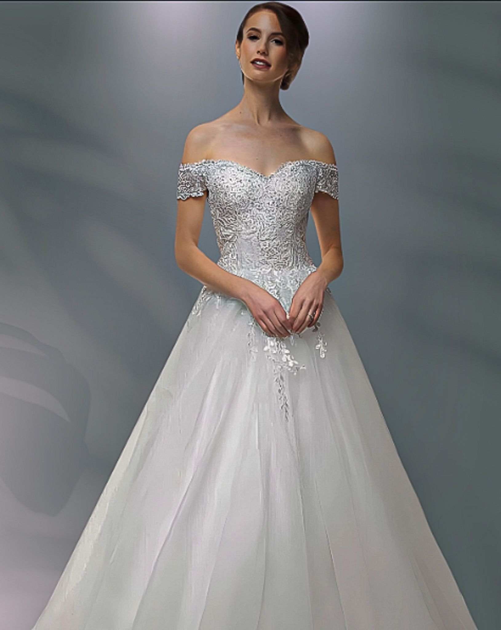 Woman in a white off-the-shoulder wedding gown with a full skirt, posing against a gray background.