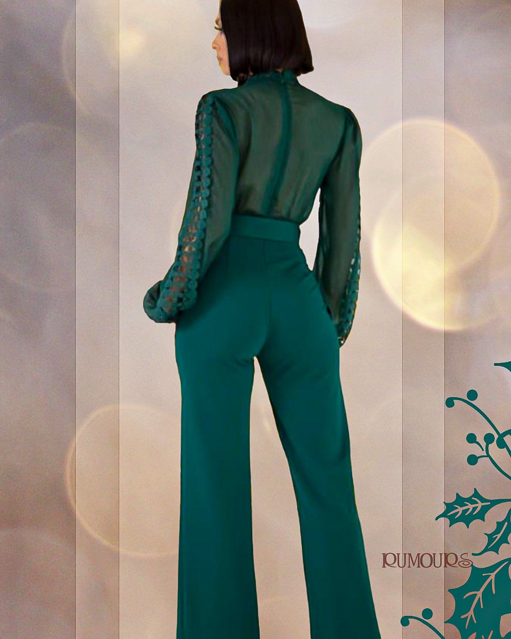 Woman in green jumpsuit with sheer sleeves and high neck, posing from behind.