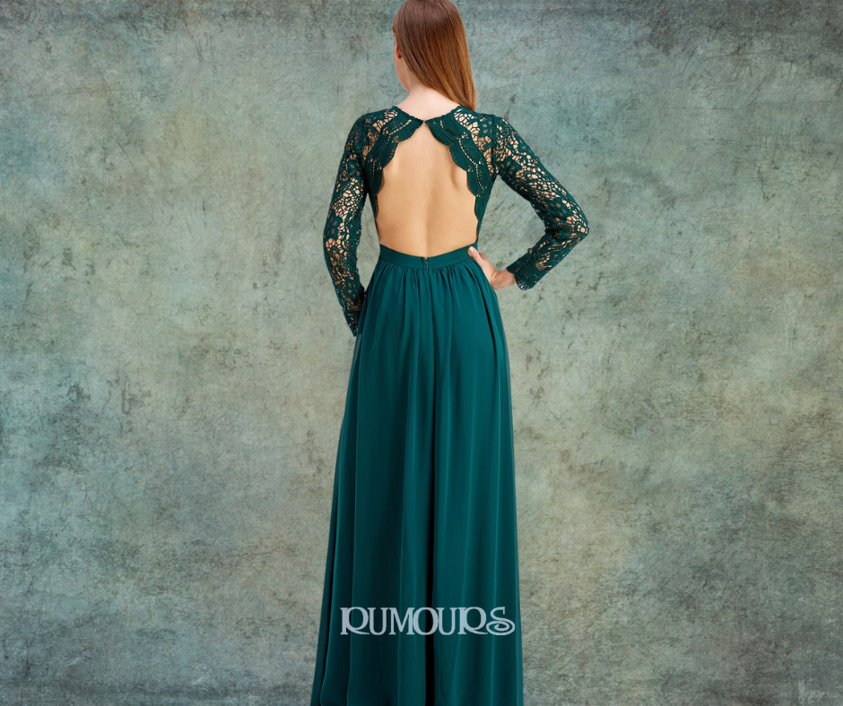 Woman in a teal, open-back gown with lace sleeves, standing against a textured gray wall.