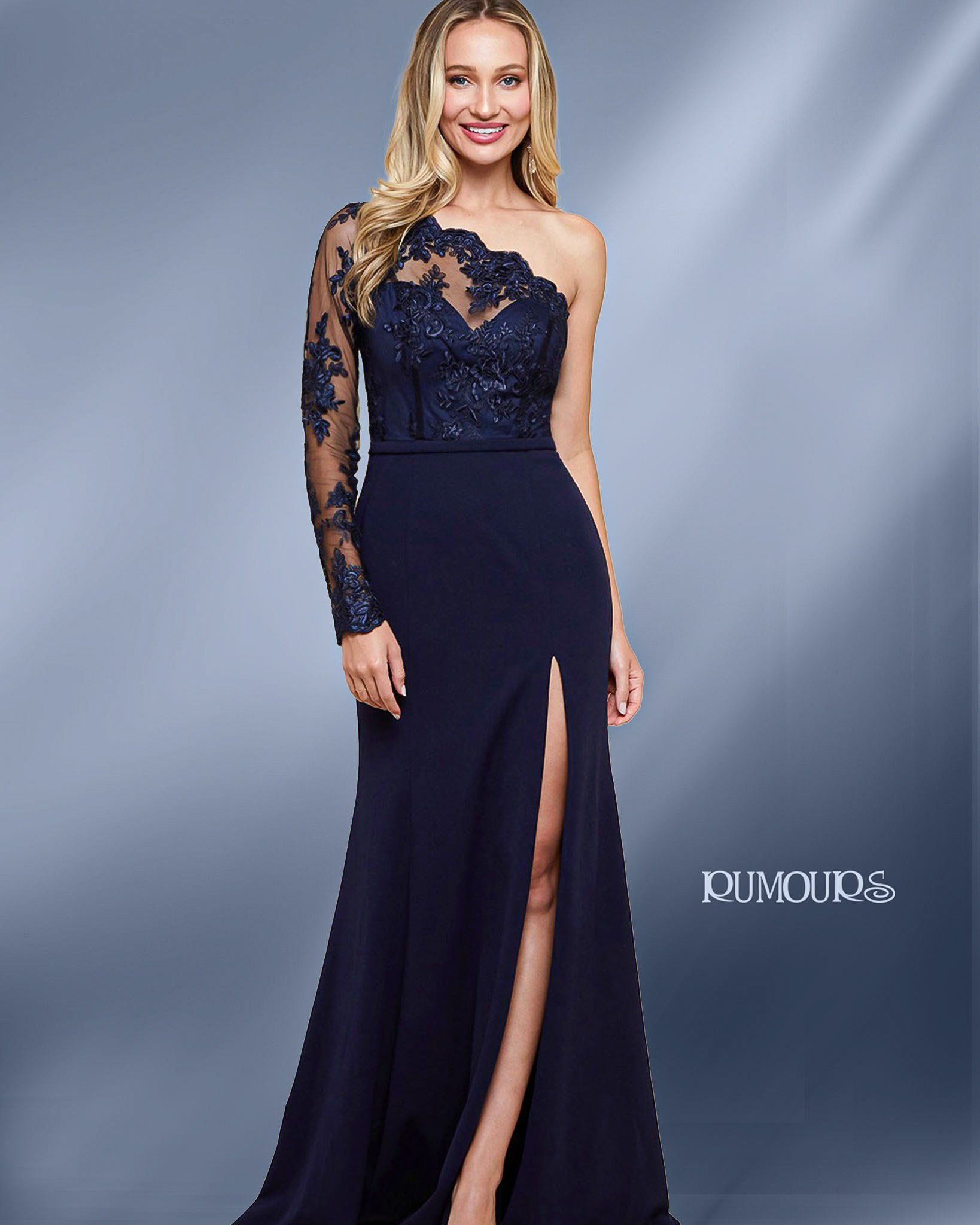 Woman in navy one-shoulder gown with sheer lace sleeve and thigh slit, posing in a studio.