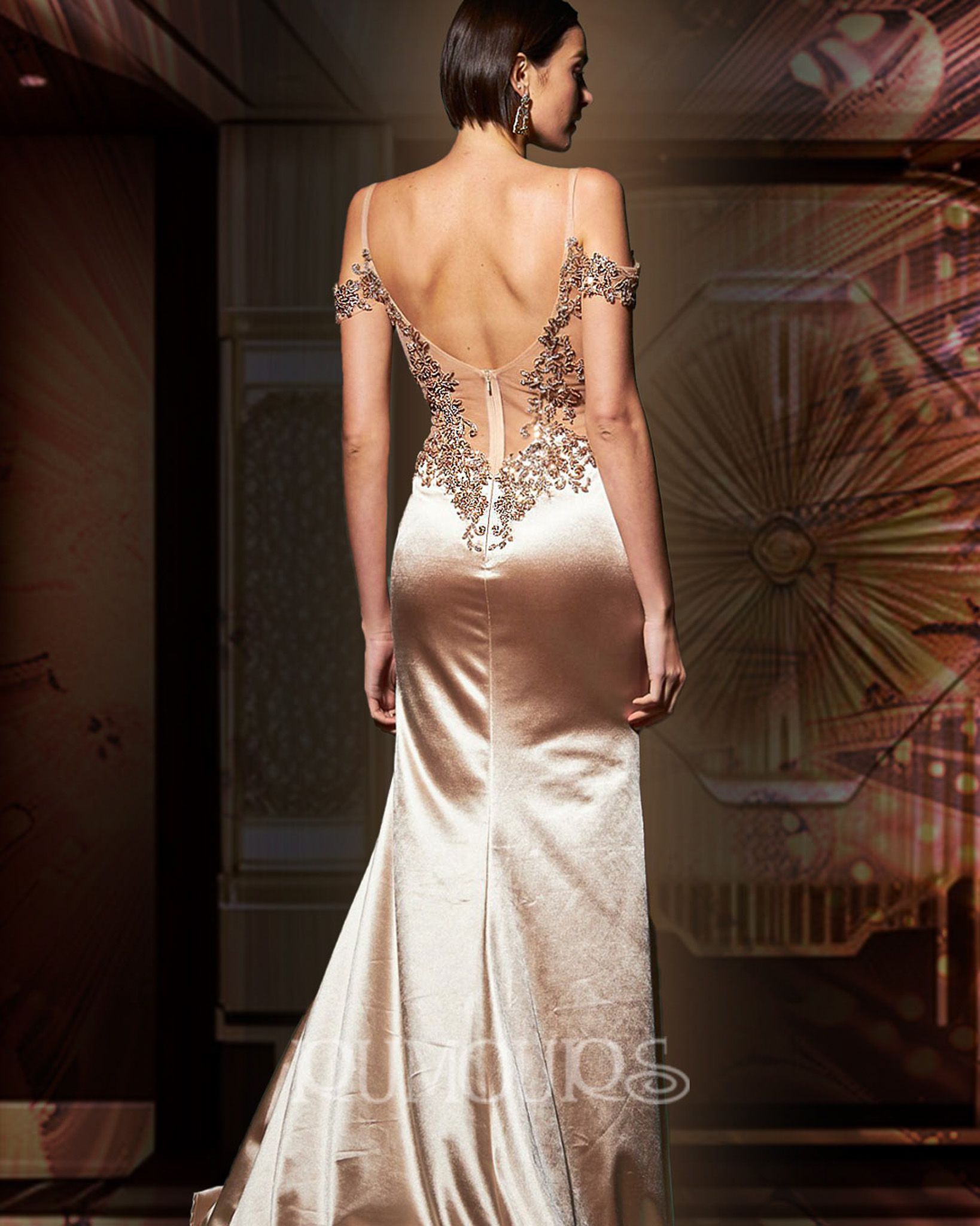 Woman in a backless, champagne-colored satin gown with decorative back detailing, standing in front of a patterned backdrop.