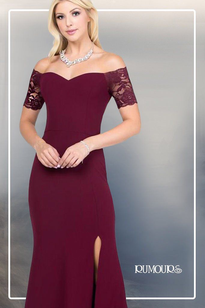 Woman in a burgundy off-the-shoulder dress with lace sleeves and a thigh slit, wearing a necklace, holding a small clutch.