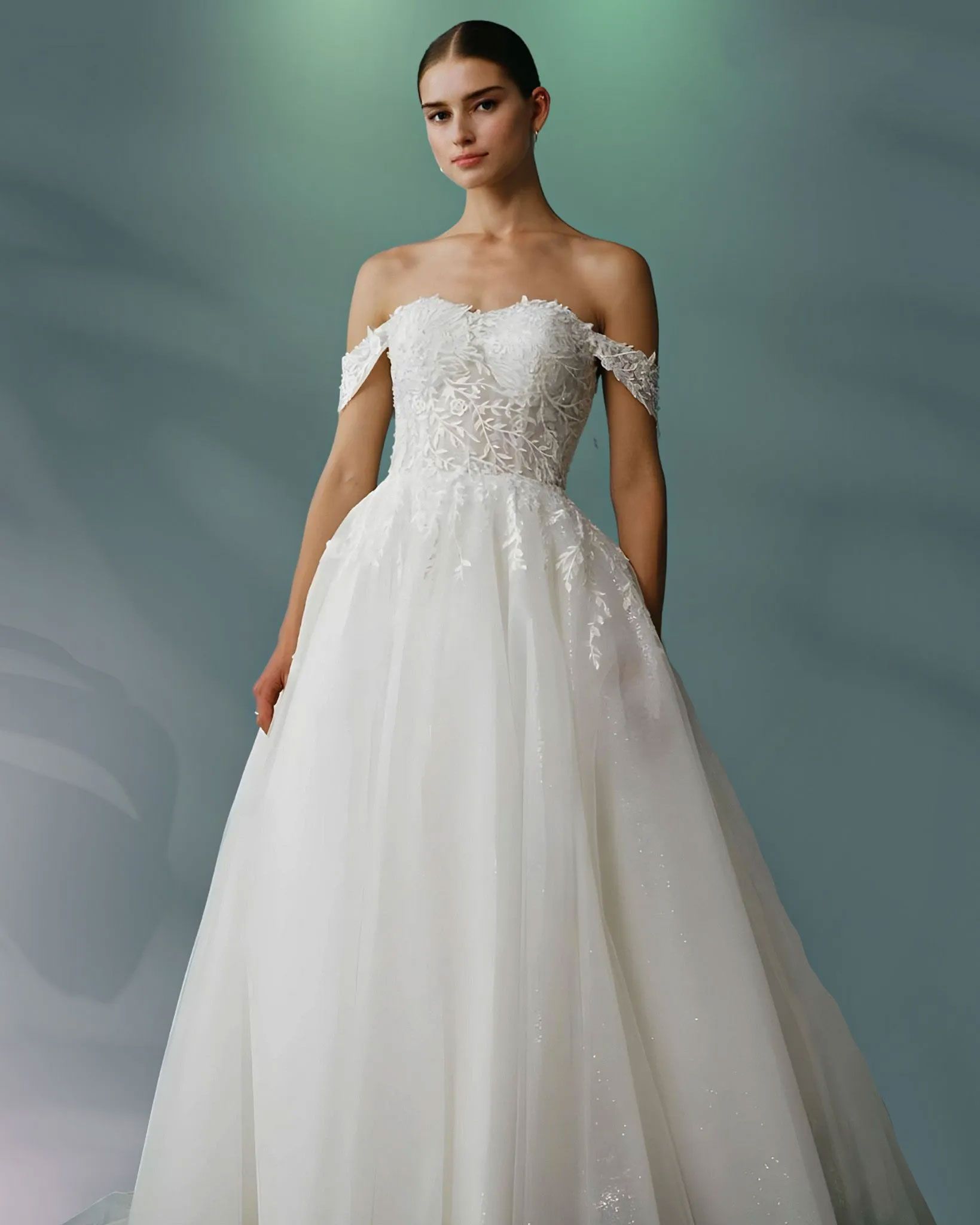 Woman in off-the-shoulder wedding gown with floral bodice, full skirt, standing against a blurred green backdrop.