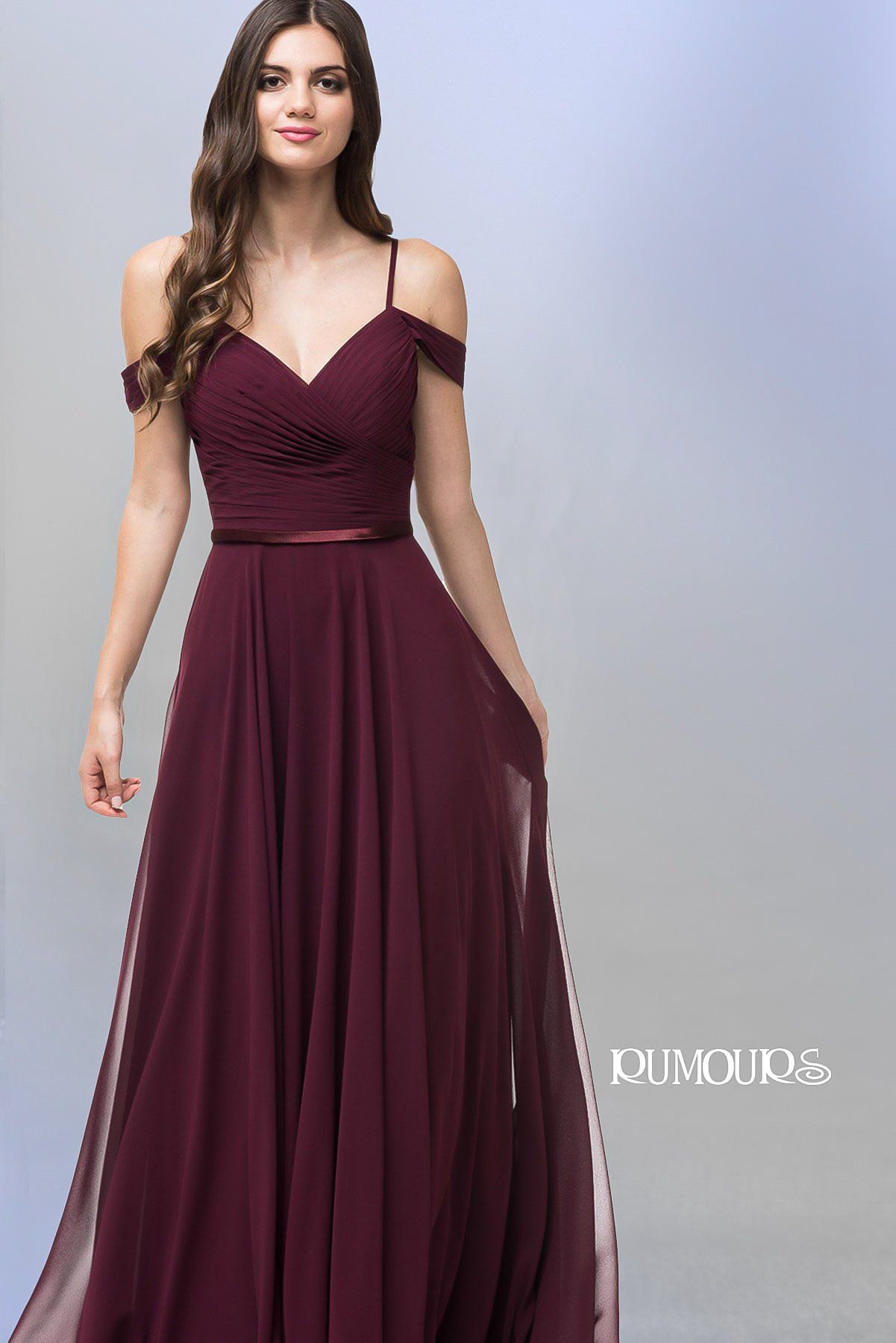 Woman in a burgundy off-the-shoulder formal dress with flowing skirt, standing in a studio setting.