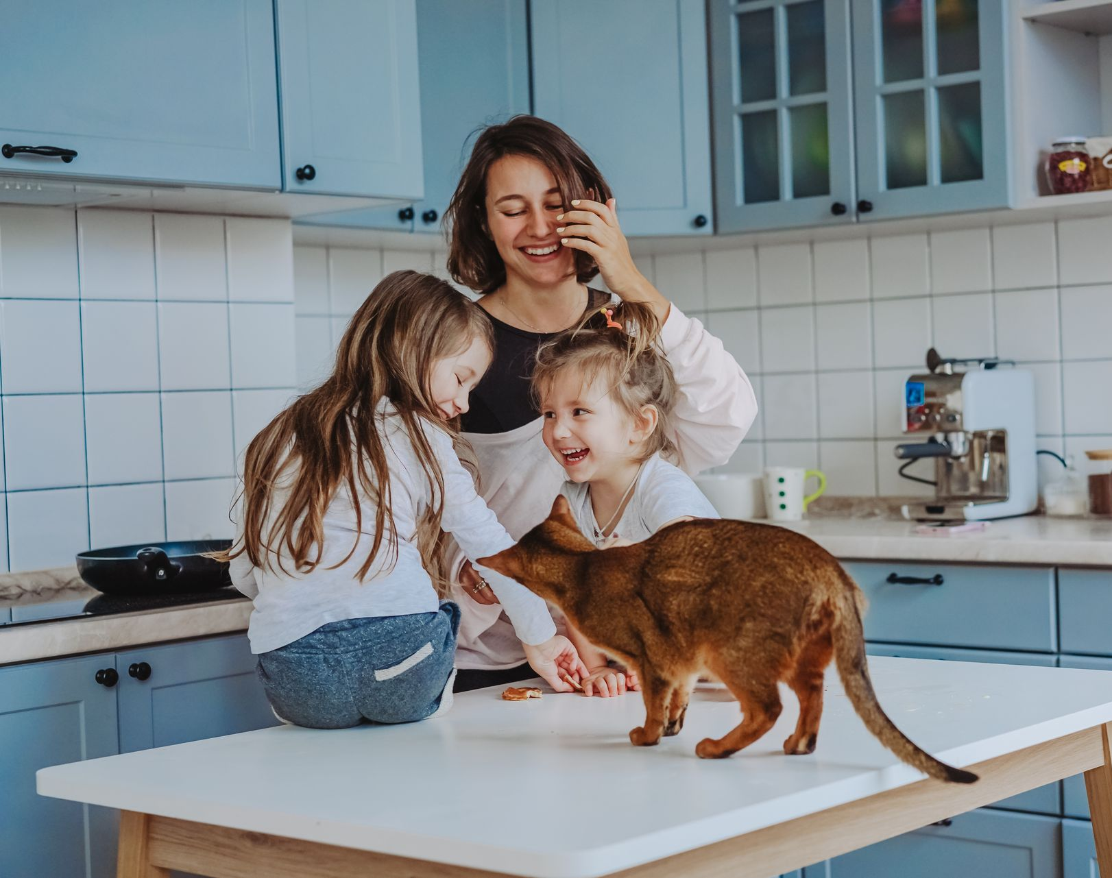 Woman in kitchen on phone, smiles at two girls and cat on table. Blue cabinets.