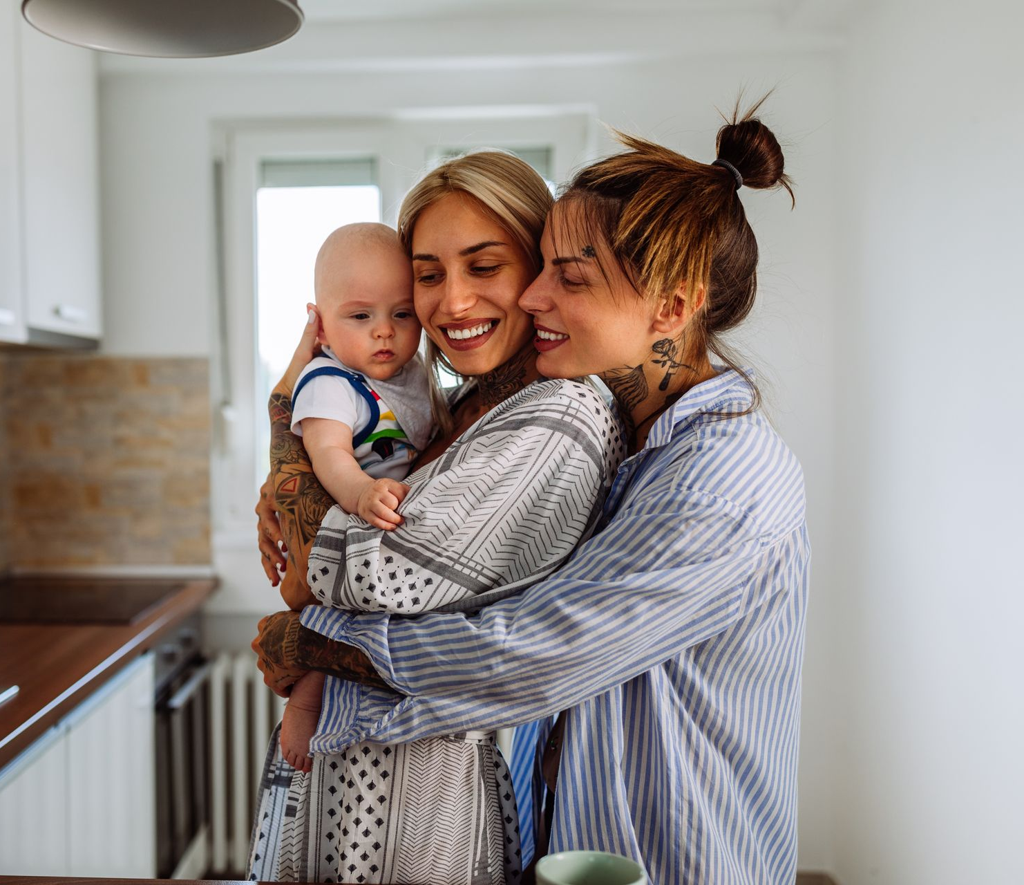 Two women embracing a baby in a kitchen. One woman holds the baby while the other hugs them, smiling.