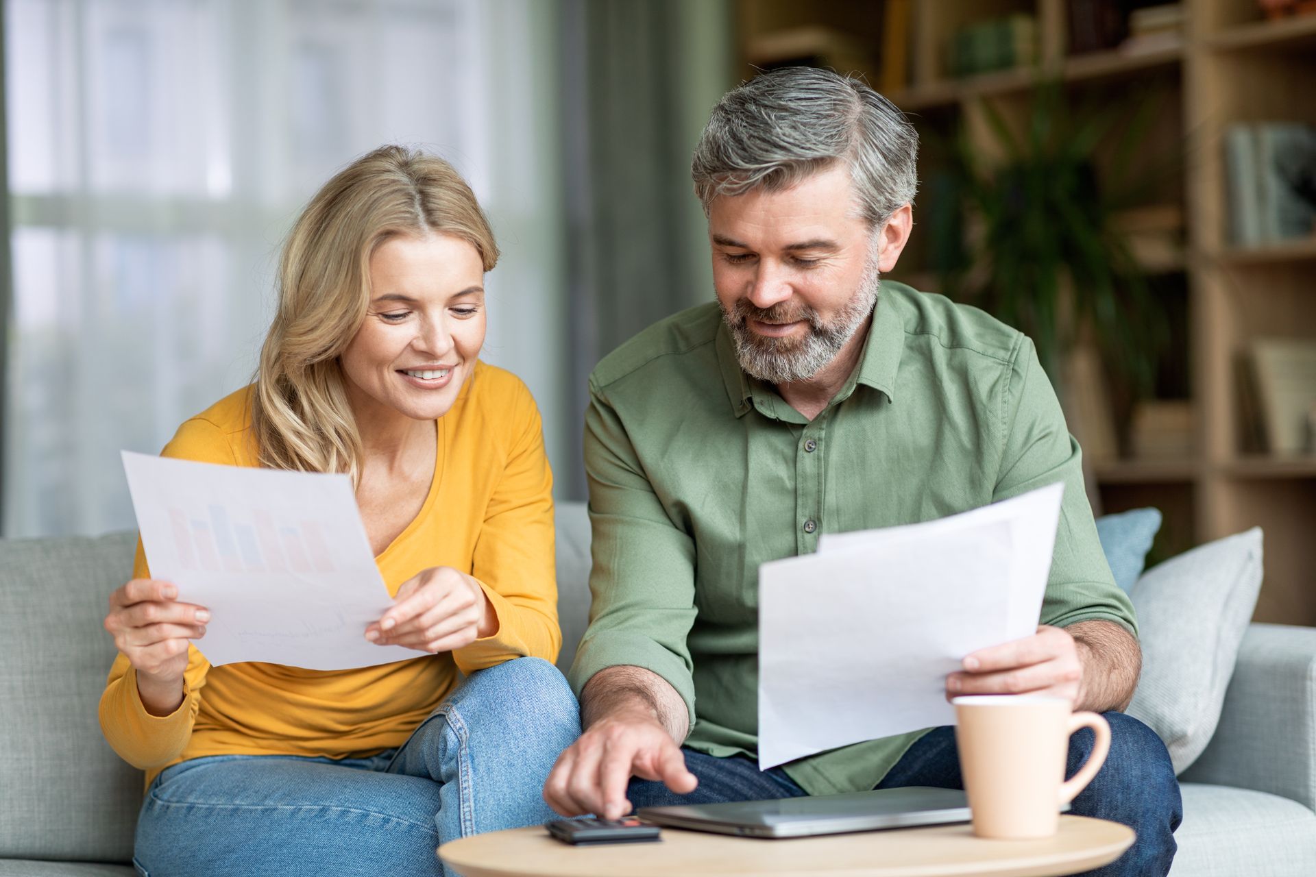 Couple reviewing documents, using a calculator, smiling, sitting on a couch. Living room setting.