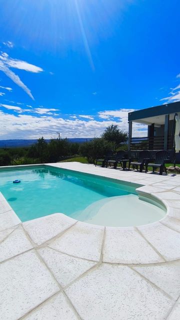 Piscina de color turquesa brillante junto a un patio moderno bajo un cielo azul claro con montañas en la distancia.