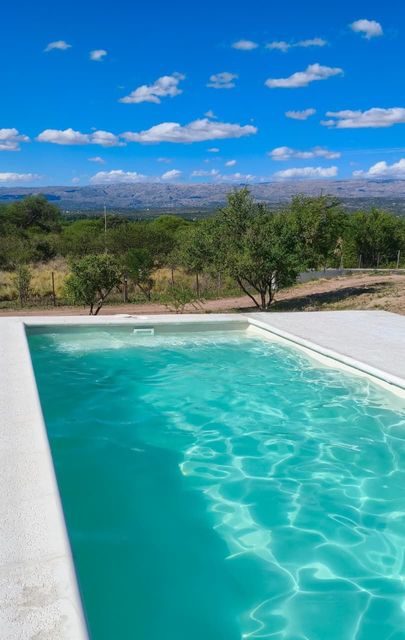 Un estanque turquesa junto a vegetación seca y montañas distantes bajo un cielo azul brillante.