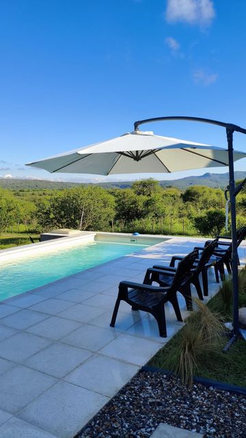 Patio junto a la piscina con sombrilla blanca, sillas negras y vistas a la montaña bajo un cielo azul.