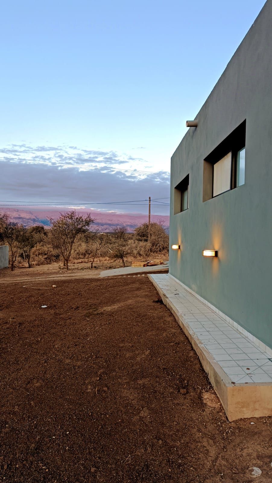 Exterior de una casa moderna con pared azul, pequeñas ventanas iluminadas y un patio de tierra con vistas a un cielo rosado al atardecer.