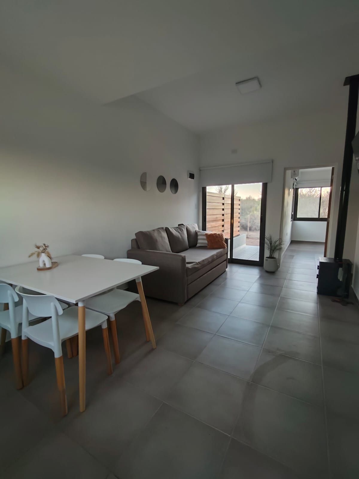 Luminoso salón con sofá gris, mesa de comedor pequeña, suelo de baldosas y puerta corredera de cristal que da al balcón.