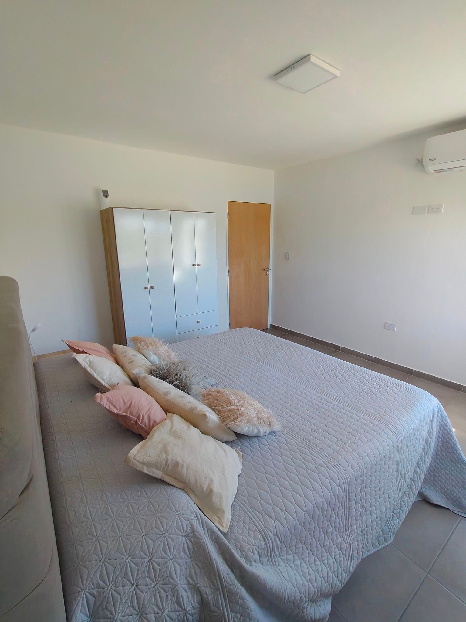 Dormitorio con colcha gris, almohadas, armario blanco y suelo de baldosas en una habitación luminosa.
