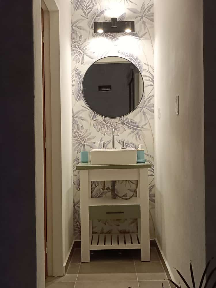 Mueble de baño estrecho para pasillo con espejo redondo, papel pintado estampado y lavabo blanco.