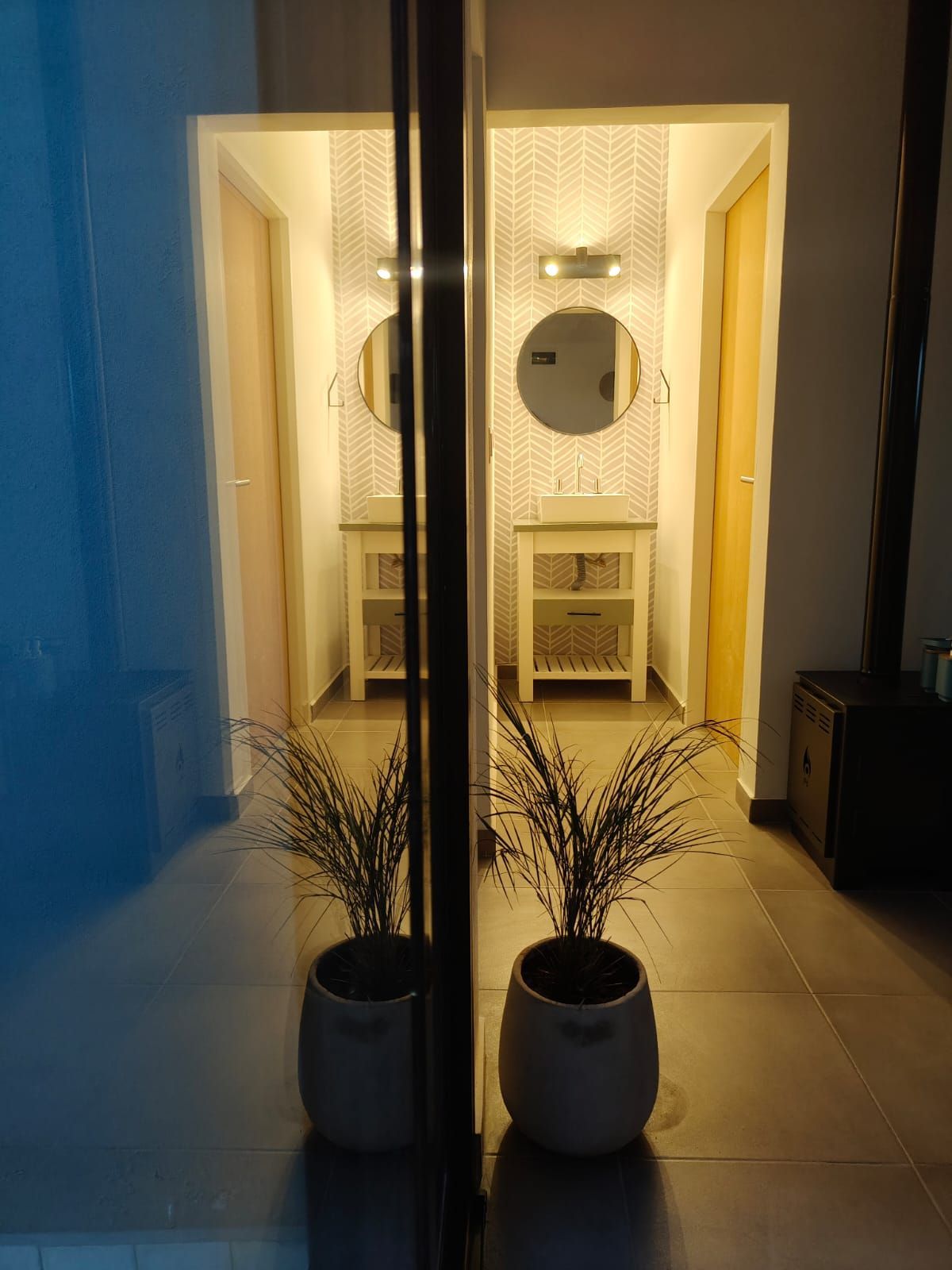 Dos macetas frente a una puerta iluminada que conduce a un pequeño baño con espejos.