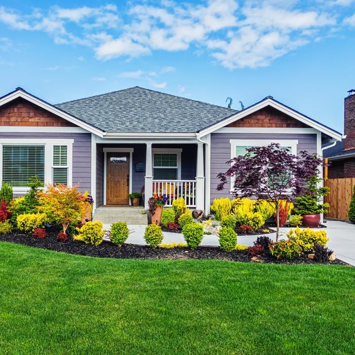 Single-story gray house with white trim, front porch, landscaped garden, and green lawn under a blue sky