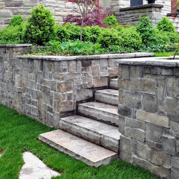 Stone steps leading to a raised stone planter with green shrubs beside a brick house.