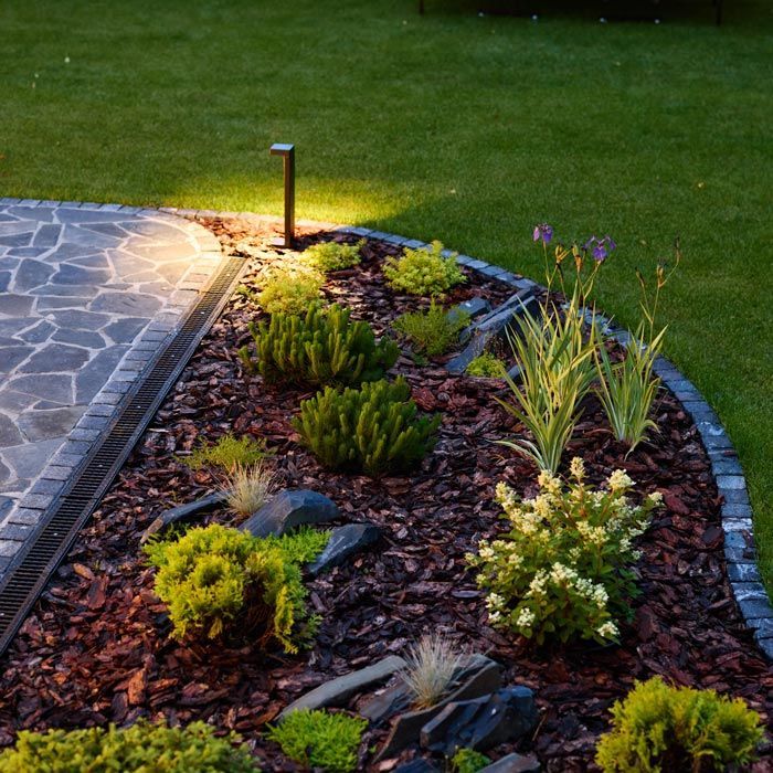 Curved garden bed with mulch, colorful plants, and a stone patio edge at dusk